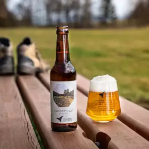 Wanderlust Bier steht auf Bierbank mit Wanderschuhen im Hintergrund Wiese und Bäume unscharf
