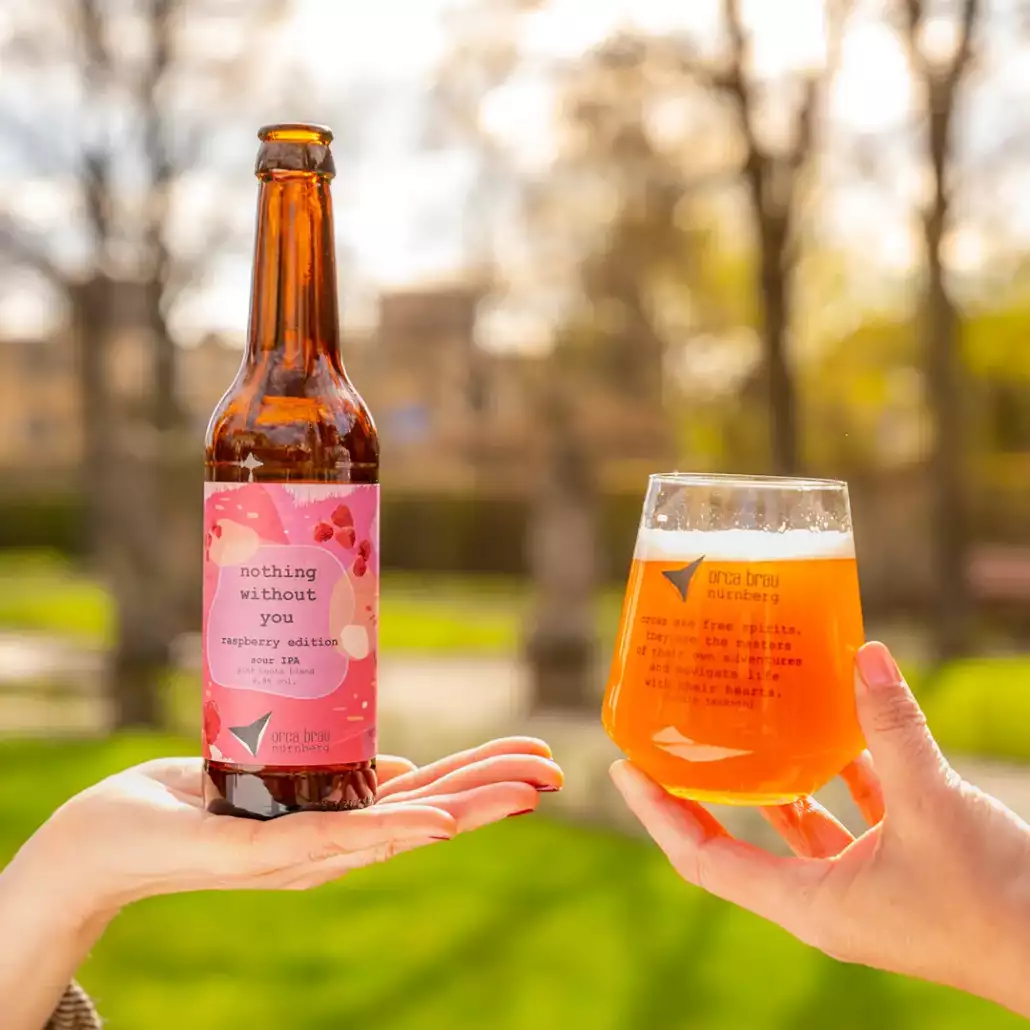 Flasche Bier auf einer ausgestreckten Frauen Hand mit Glas Bier in einer Frauenhand daneben, in Hintergrund unscharf ein Park im Frühling